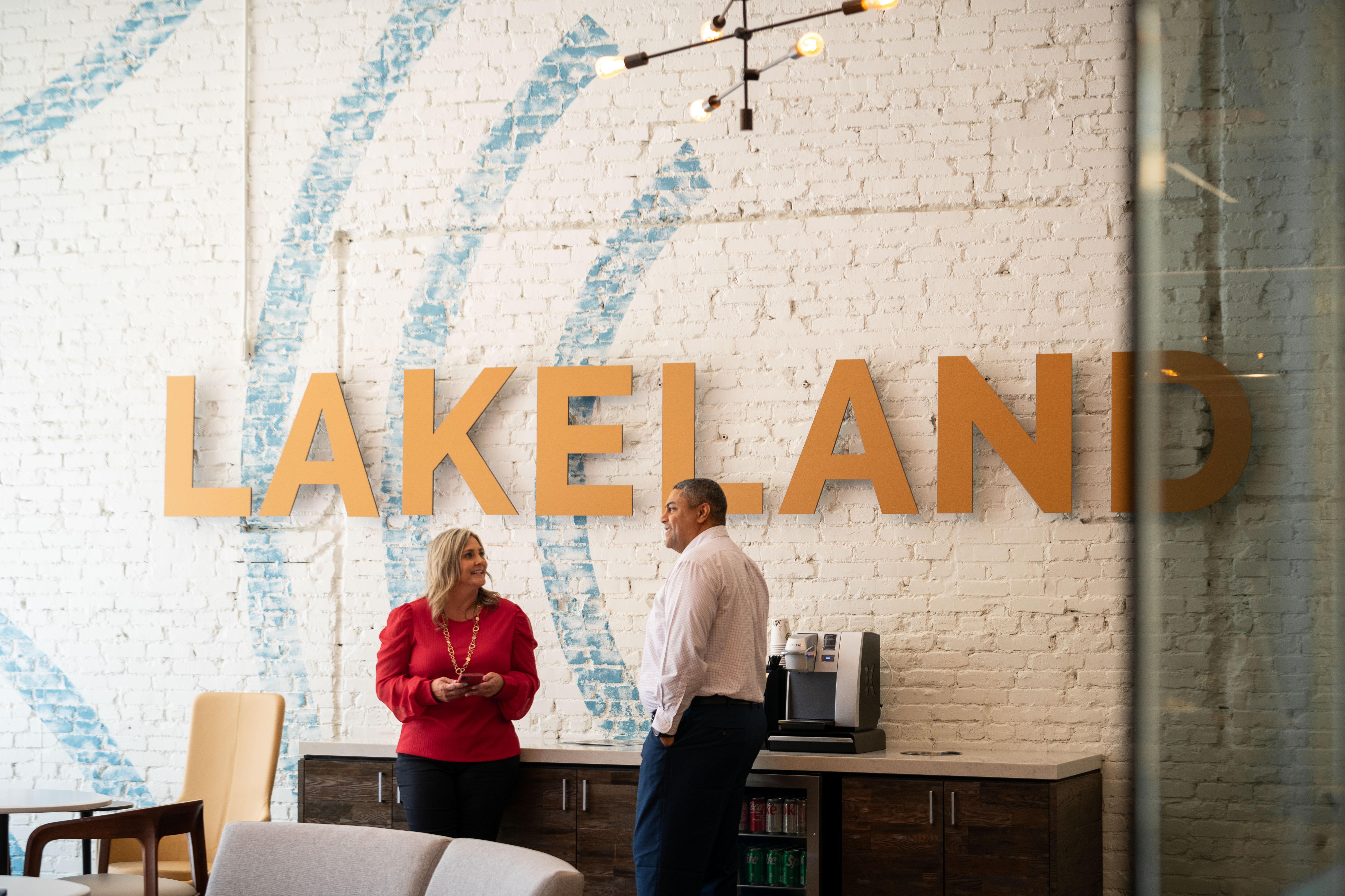 Young White woman in a red top and Hispanic gentleman standing in front of a coffee station in front of a white painted brick wall with the word LAKELAND in yellow letters, in a Crews Bank & Trust, having a discussion