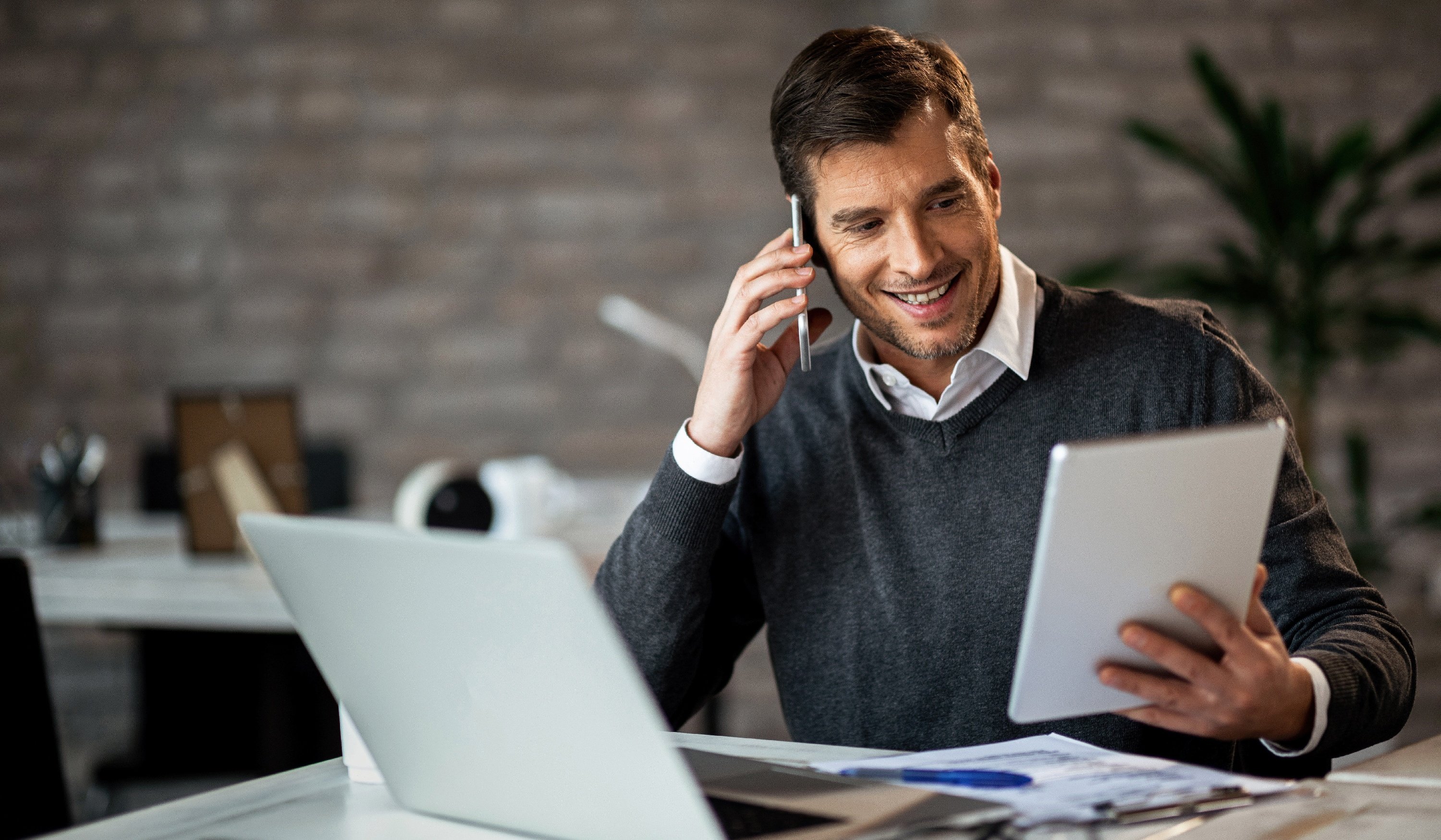 Middle-aged white male with computer in front of him and holding a loan application, talking on a cell phone.