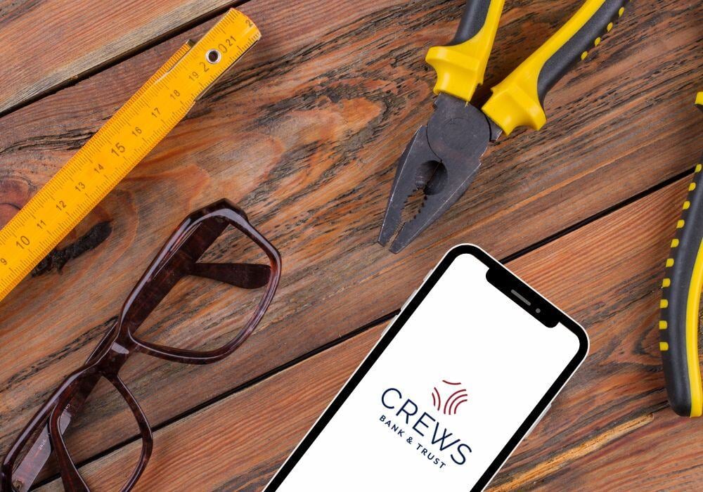 eye glasses, tape measure, pliers, cellphone with Crews Bank logo all on a wooden floor or tabletop