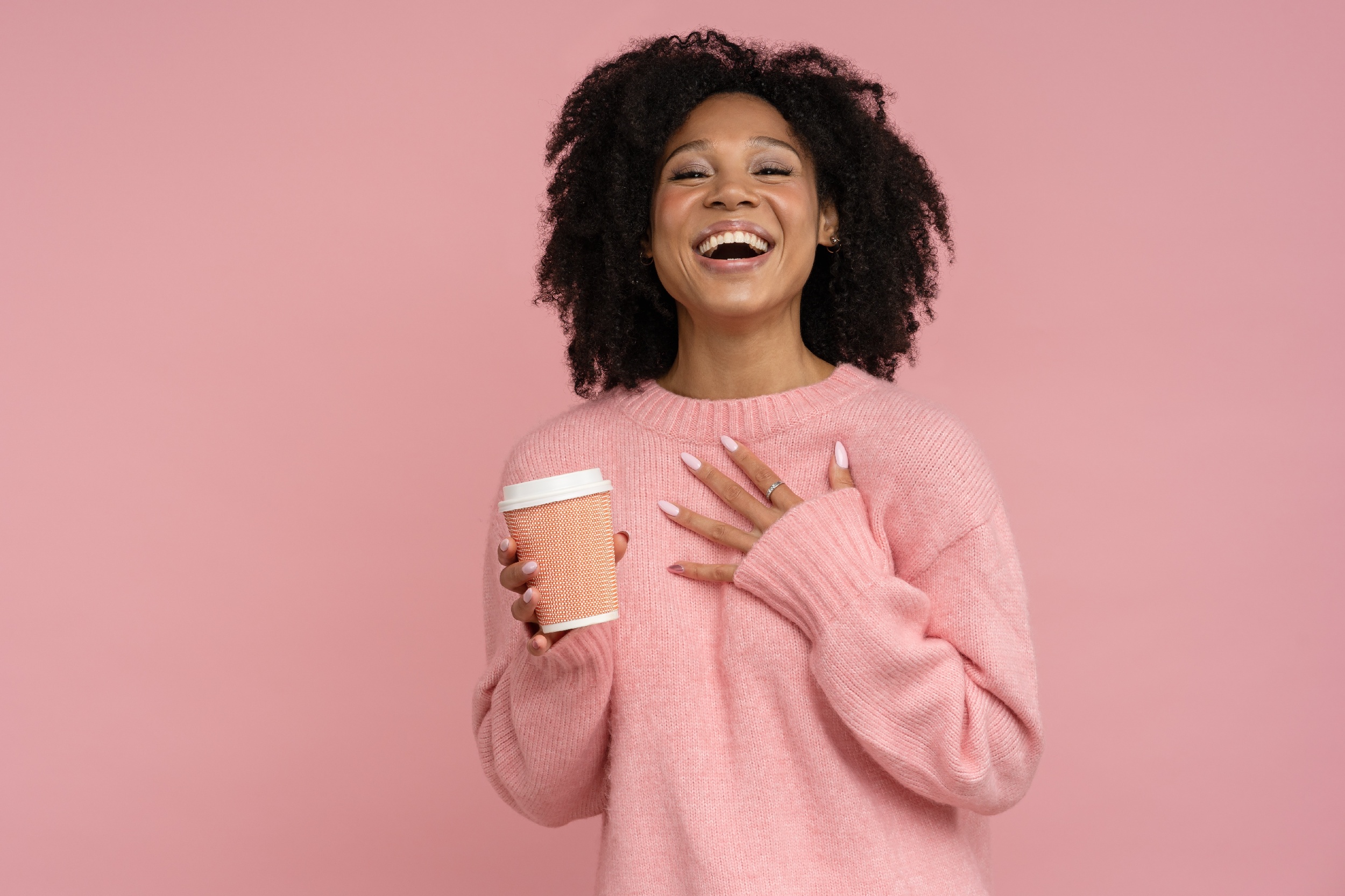 Smiling young black woman in a pink sweater holding a take-away coffee cup, against a pink background