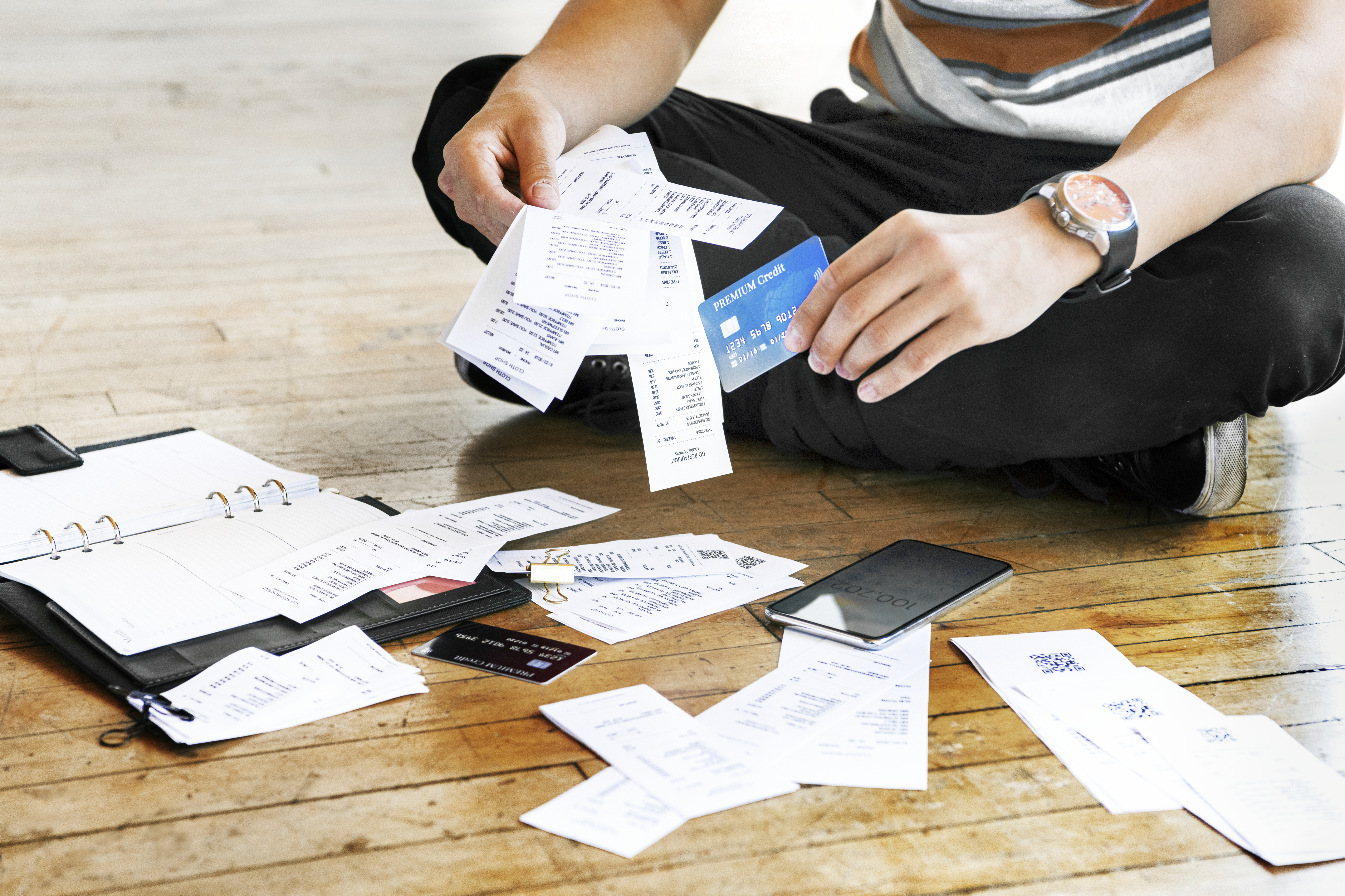 Young man sitting on wood floor with many receipts spread out around him, looking at phone