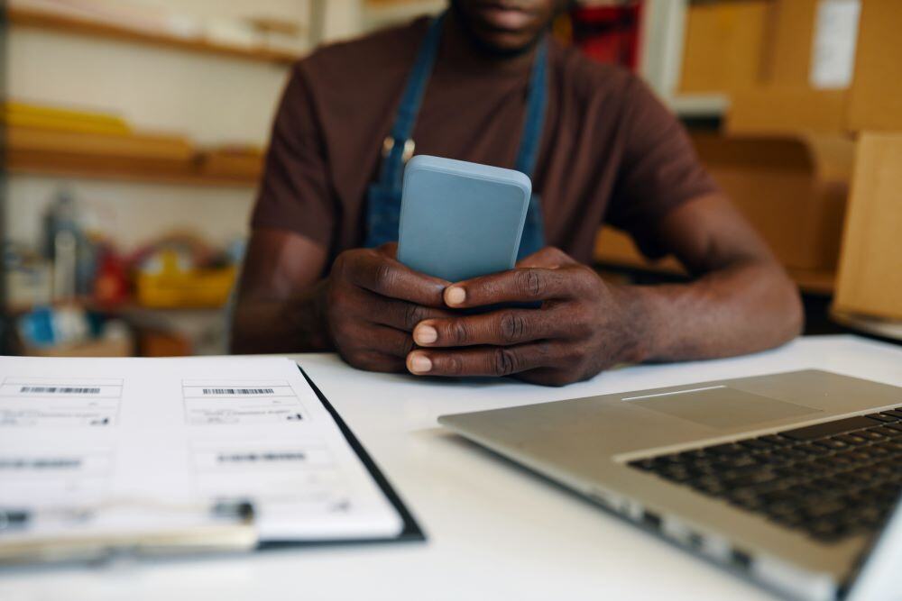 Yound black entrepreneur sitting at a desk, looking at his phne, with a computer and a binder on the desk with him