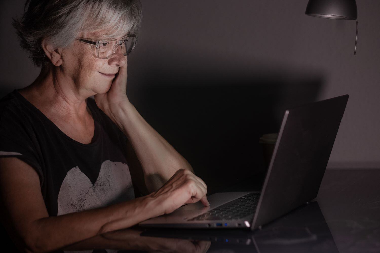 Older woman sitting at a desk, in a dark room, working on a laptop.