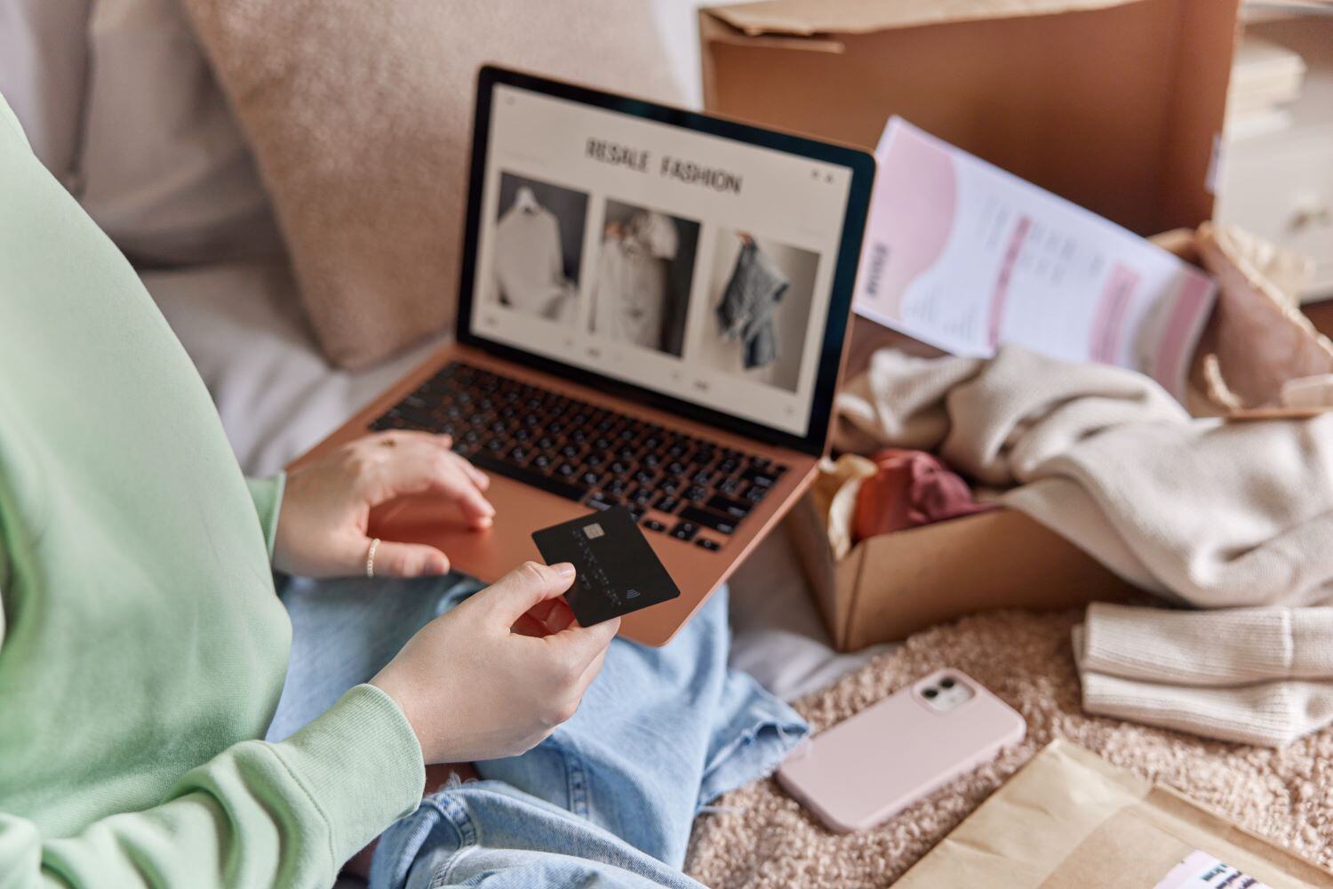 View of a young woman's hands holding a credit card and sitting cross-legged with a laptop on her lap, open to a shopping website.