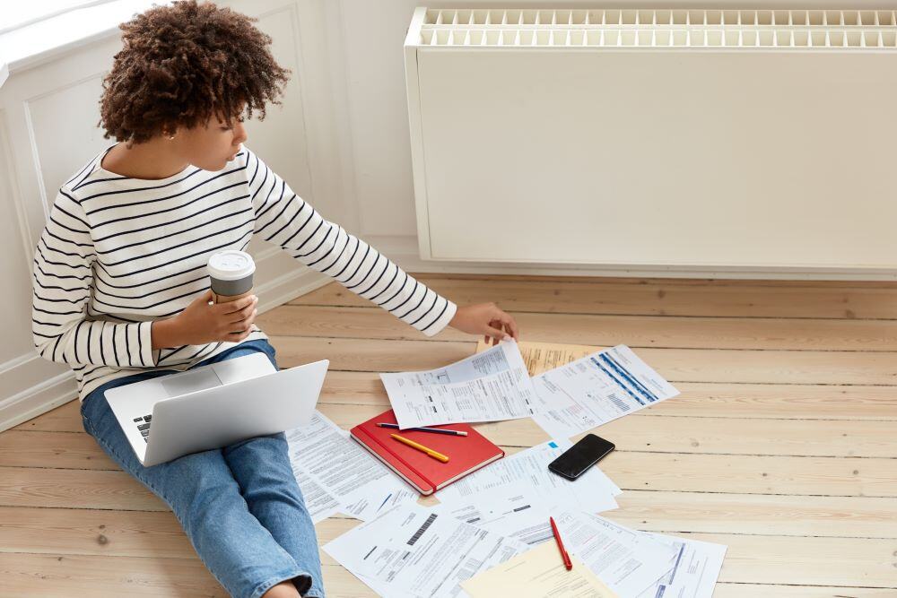 Young African American woman seated on a wooden floor, holding a take-out coffee, with a laptop on her lap, surrounded by bills strewn about her on the floor. 